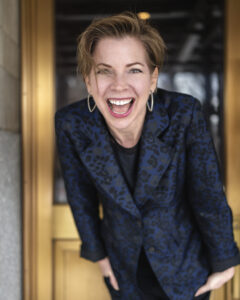 Headshot of Kate O'Neill shows a person laughing joyfully while leaning forward, wearing a stylish blue patterned blazer, standing in front of a golden door.